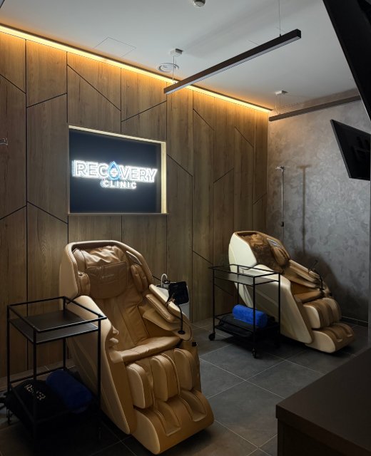 Lounge kliniky – dve béžové masážne kreslá pri drevenej stene s podsvieteným logom Recovery Clinic a stojanmi na infúzie